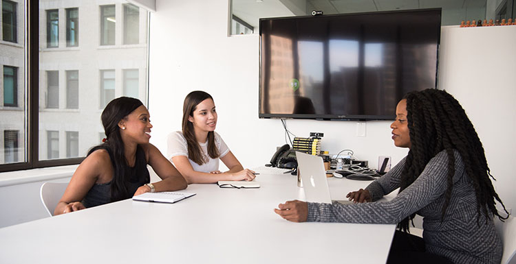 Three people are seated and talk at a table in a conference room.