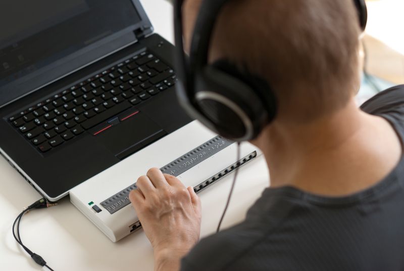 Man using a computer with headphones and a refreshable Braille display