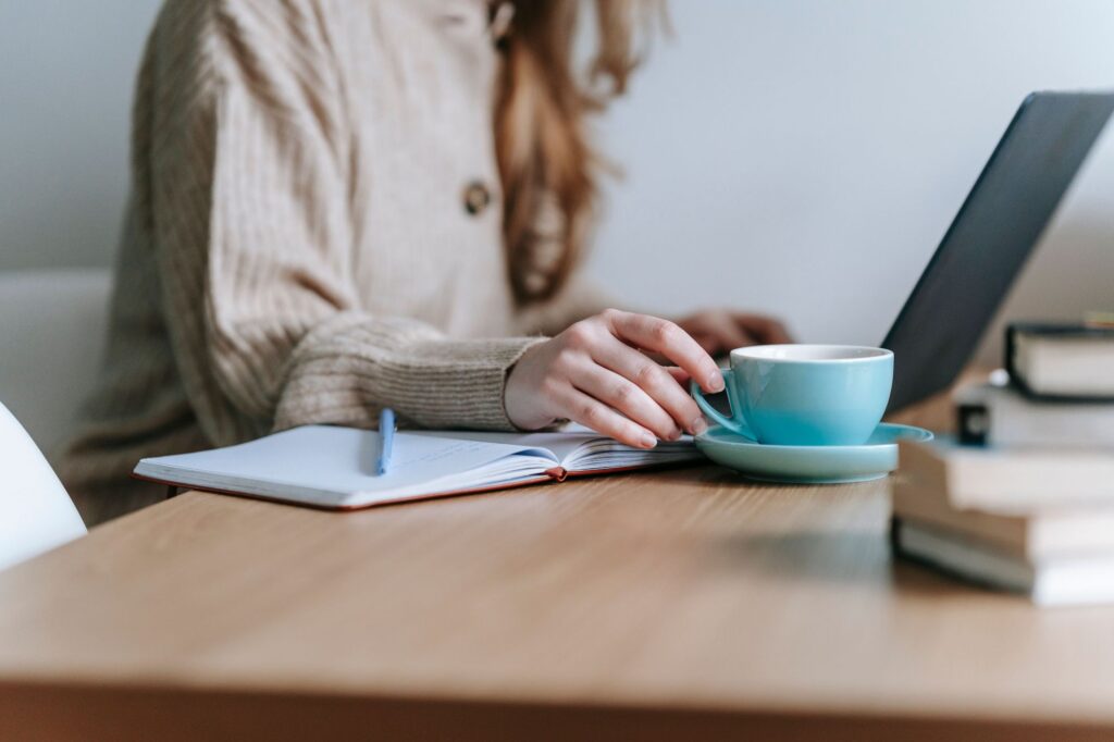 A women shown from the shoulders down drinking coffee and typing on a laptop with a notebook and pen nearby
